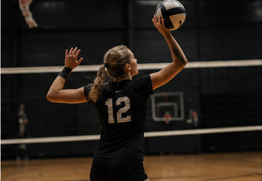 A volleyball player setting the ball, back to camera, basketball hoop visible in the gym behind her.