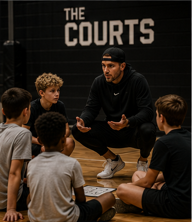 A coach crouching to talk with his team courtside, with THE COURTS lettering on the wall behind them.